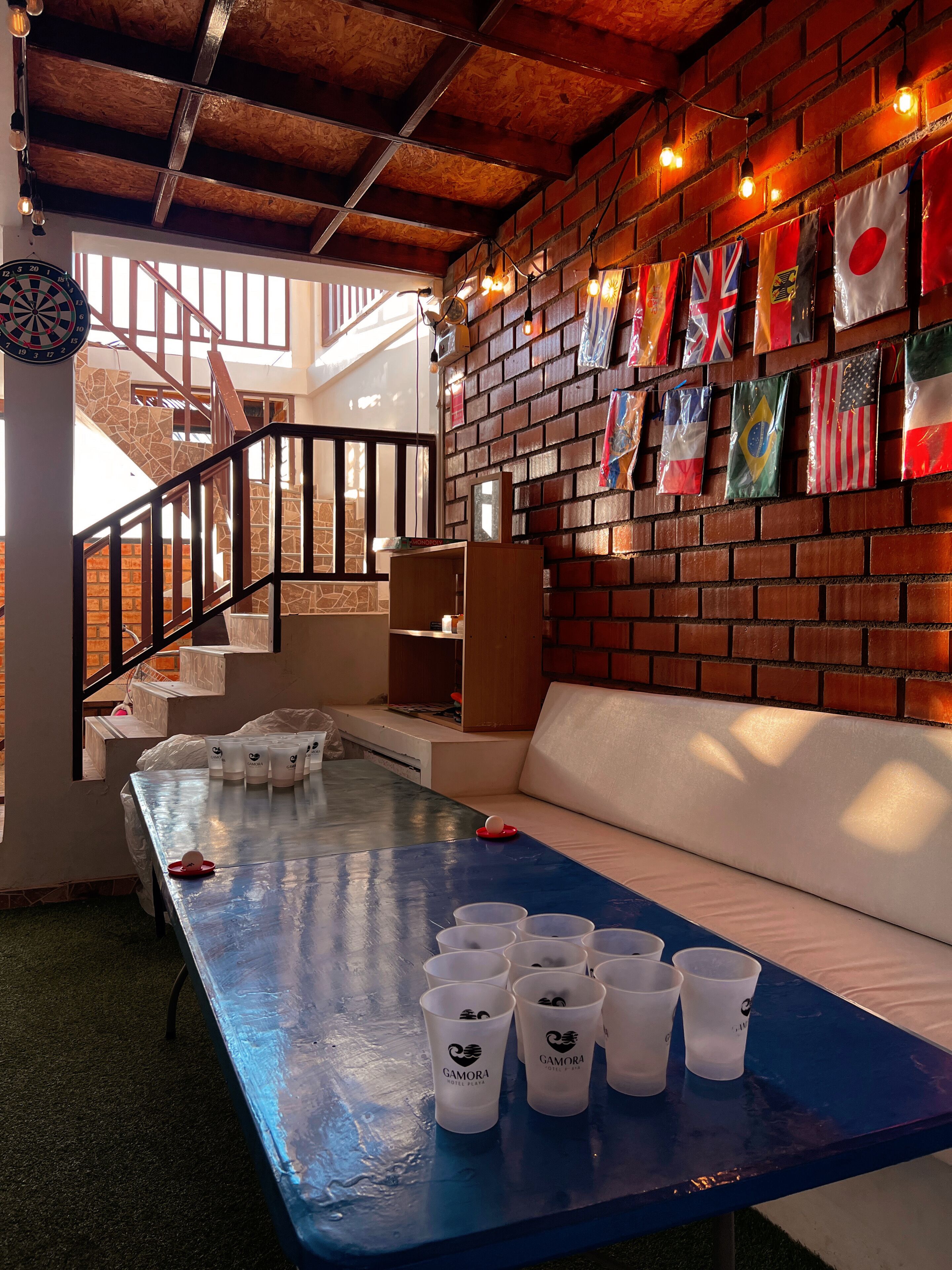 A room with a blue table, white cups, a dartboard, and a wall decorated with various flags.