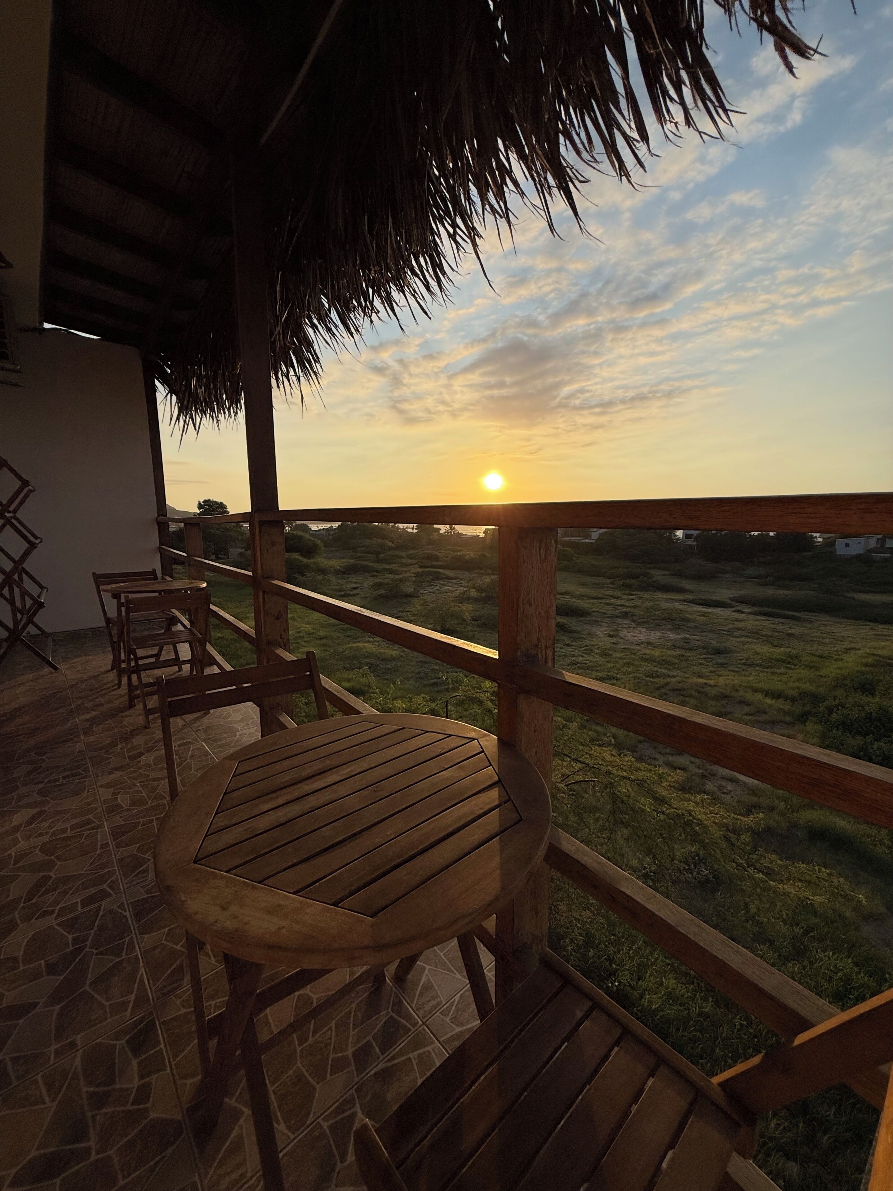 Una terraza de madera con una mesa y sillas, un techo de paja y vista a un campo de hierba al atardecer.