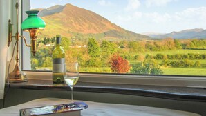 A table with a bottle of wine, a glass, and a book, with a view of a mountainous landscape through a window. - 2 bedroom accommodation in Bassenthwaite, near Keswick (Bassenthwaite, near Keswick)