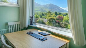 A room with a wooden table, a white chair, and a view of a green landscape with trees and mountains. - 2 bedroom accommodation in Bassenthwaite, near Keswick (Bassenthwaite, near Keswick)