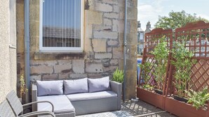 A stone building with a window, a wicker sofa, a chair, and a wooden fence with plants. - 2 bedroom accommodation in Ardrossan, near Ayr (Ardrossan, near Ayr)
