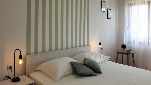 A neatly made bed with white and green pillows, a bedside lamp, and a small table with a plant and a bag. - Apartment with balcony, parking near the center and the sea (Crikvenica)
