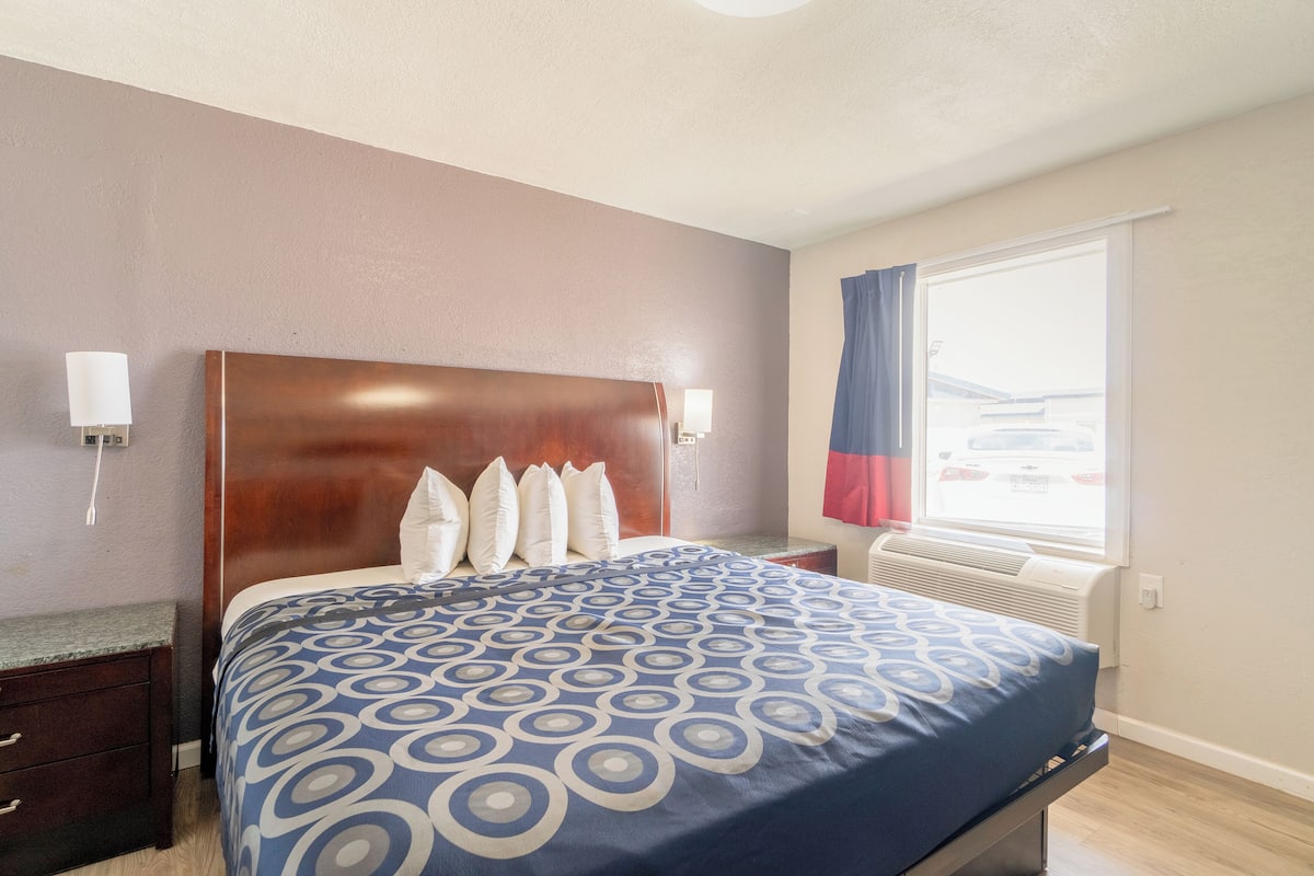 Een hotelkamer met een groot bed, een houten hoofdeinde, een nachtkastje, een lamp, een raam met gordijnen en een airconditioning.