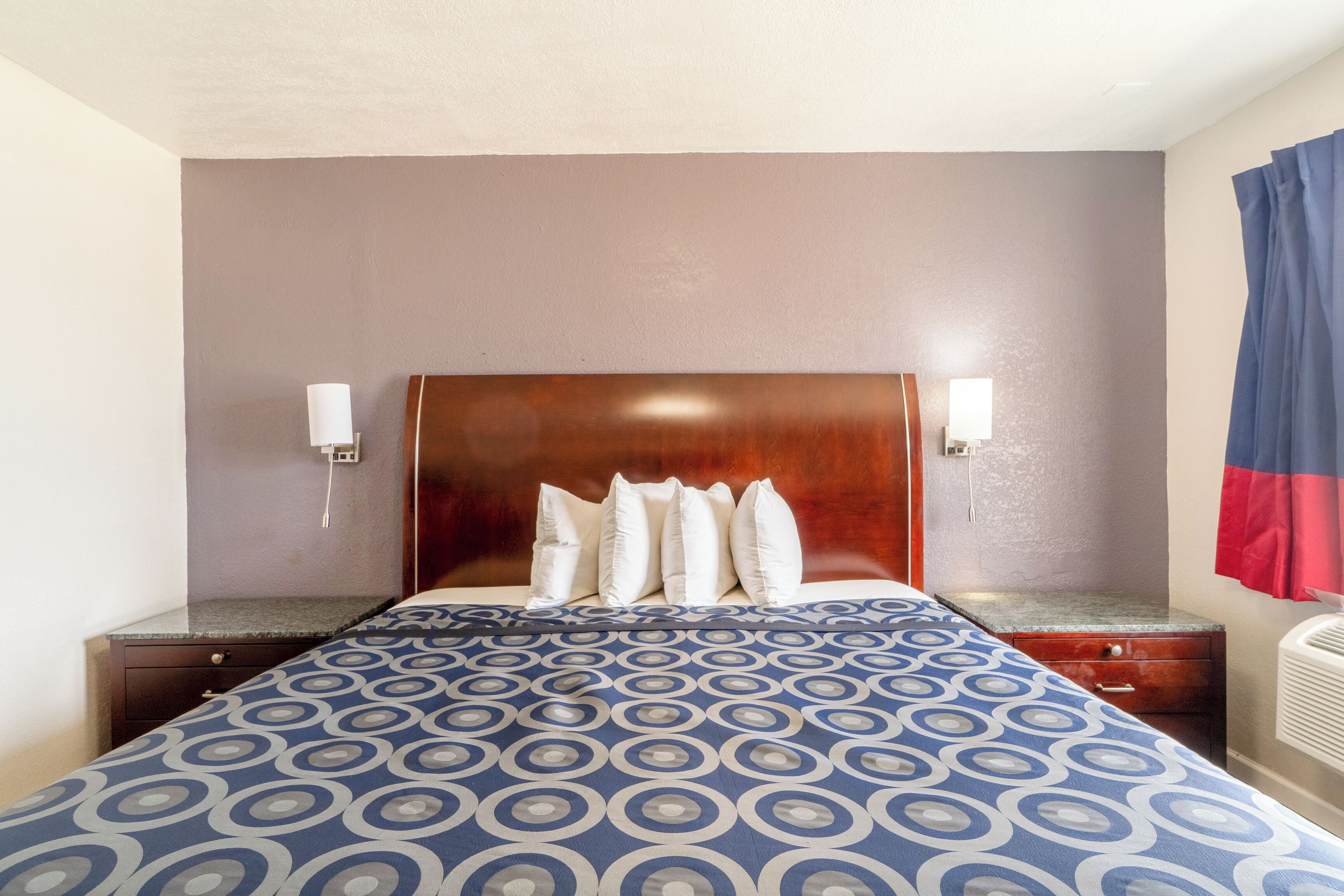 A hotel room with a wooden headboard, a patterned bedspread, a nightstand with a lamp, and a wall-mounted heater.
