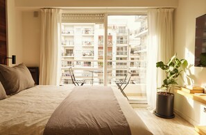 A hotel room with a bed, a potted plant, a balcony with a table and chairs, and a view of a residential building. - Hestia at Cabrera 3200 (Buenos Aires)