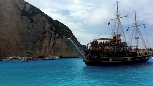 Ein großes Segelschiff liegt vor einer felsigen Küste mit klarem, blauem Wasser im Hafen.