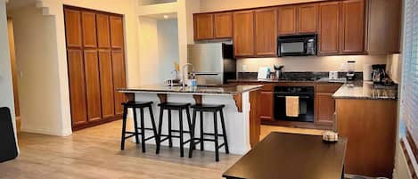 A modern kitchen with wooden cabinets, a central island with stools, a built-in oven, and a microwave.
