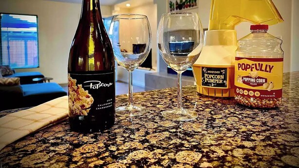 Una bottiglia di Popcorn Chardonnay, due bicchieri da vino, una macchina per popcorn e una bottiglia di Popfully granella di mais che scoppietta su un bancone della cucina.