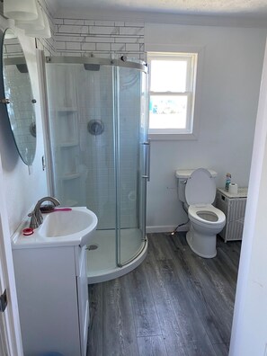 Een badkamer met een douchecabine van glas, een witte wastafel, een toilet en een rieten mand.