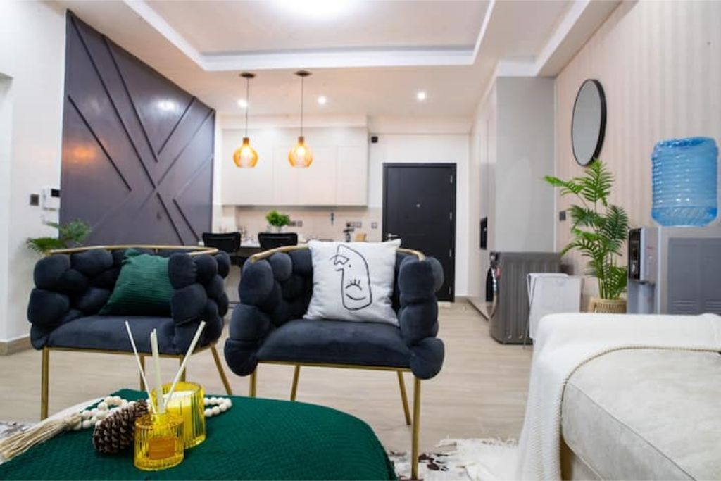 Una sala de estar moderna con dos sillones oscuros, un tapete verde y un sofá blanco. Hay dos lámparas colgantes del techo.