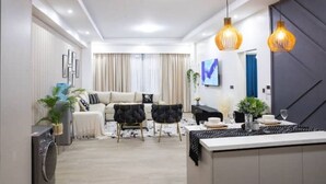 Una sala moderna con un sofá, dos sillones, una mesita de centro y un comedor con mesa y sillas. Hay lámparas colgantes sobre la mesa del comedor.