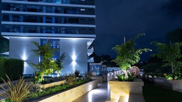 Un edificio moderno con fachada de vidrio, entrada bien iluminada y jardines ajardinados con diversas plantas y una escultura esférica.