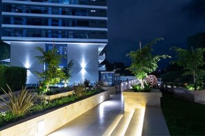 A modern building with a glass facade, a well-lit entrance, and landscaped gardens with various plants and a spherical sculpture. - 1 bedroom apartment in nairobi (Nairobi)