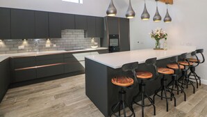 A modern kitchen with dark cabinetry, a large island with bar stools, pendant lighting, and a view of the outdoors through a window. - Langley Barn (Ashbourne)