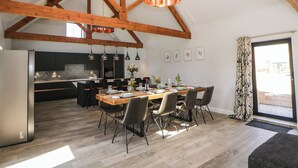 A spacious dining area with a wooden table and chairs, a kitchen in the background, and a large window with curtains. - Langley Barn (Ashbourne)