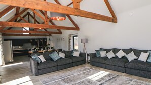 A spacious living area with high ceilings and exposed wooden beams, featuring two dark sofas with patterned cushions, a large rug, and a dining area in the background. - Langley Barn (Ashbourne)