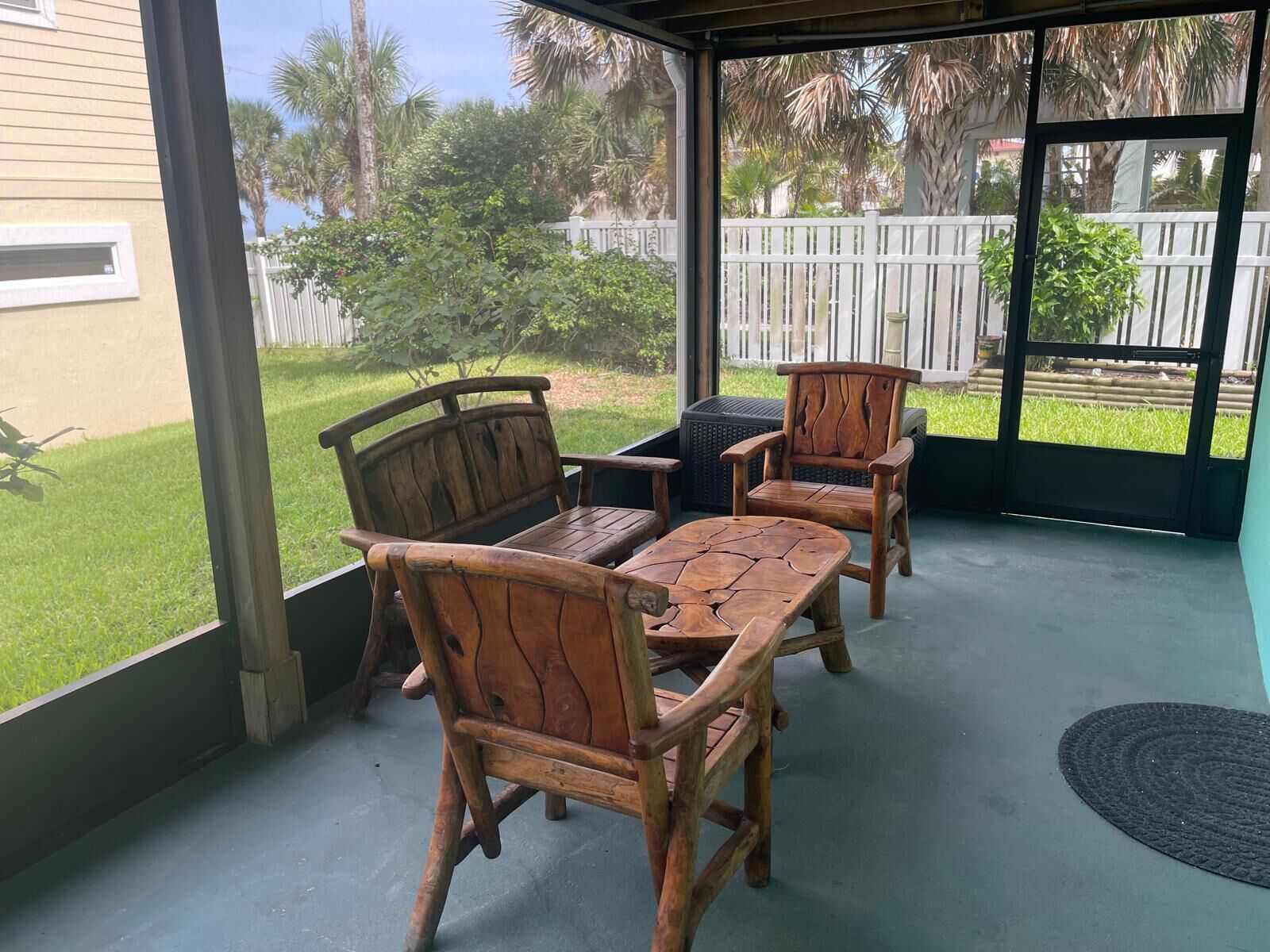 A covered patio area with wooden furniture, including two chairs and a table, and a view of a fenced yard with greenery.