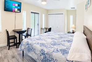 A hotel room with a bed, a desk, a chair, a TV mounted on the wall, and a door. - Seashell Studio - Private Hot Tub, King Bed, Ocean View, Short Walk to the Beach (St. Augustine)