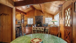 A cozy wooden cabin interior with a dining table, chairs, a fireplace, and a kitchen area. - Snowridge (Big Bear City)