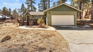 A single-story house with a garage, surrounded by a dry, grassy yard and tall pine trees. - Snowridge (Big Bear City)