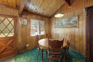 Un interior de madera con una mesa de comedor y sillas, una ventana con persianas, una puerta con vidrio tipo diamante y un cuadro colgado en la pared.