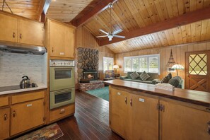 A rustic kitchen with wooden cabinets, a stone fireplace, and a ceiling fan. - Snowridge (Big Bear City)