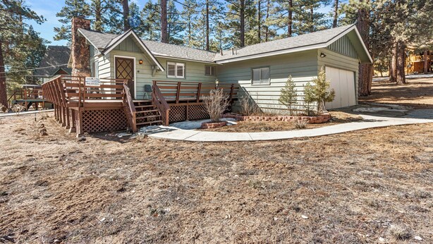 A small, single-story cabin with a chimney, a fenced porch, and a small garden area. - Snowridge (Big Bear City)