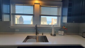 Cozinha com pia de aço inoxidável, duas janelas com persianas e vista para casas e o mar.