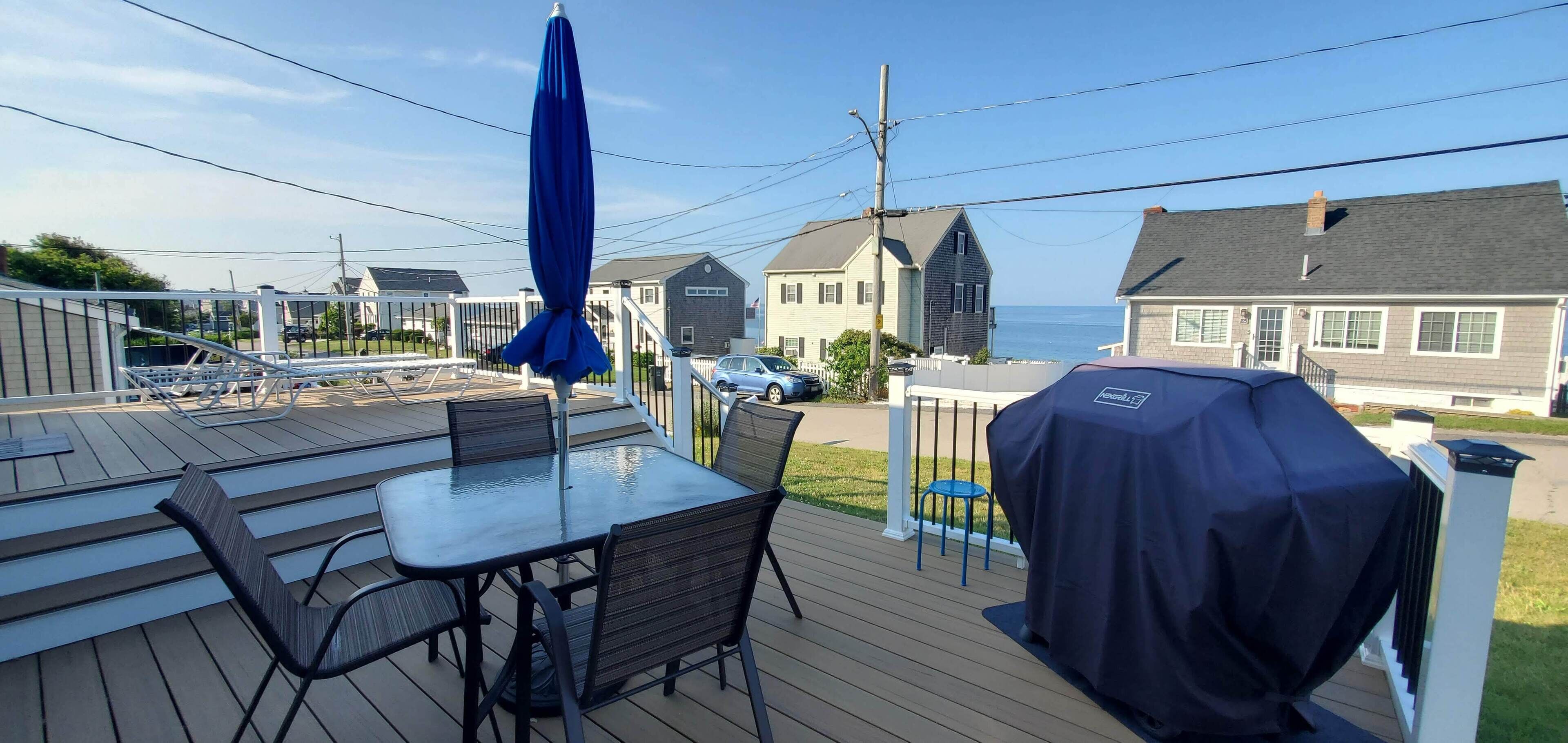 A deck with a table and chairs, a covered grill, and a view of the ocean and houses.