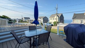 Um terraço com mesa e cadeiras, churrasqueira coberta e vista para o oceano e casas.