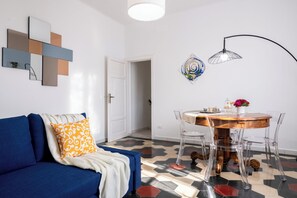A modern living room with a blue sofa, a round wooden dining table with clear chairs, and a geometric-patterned floor.