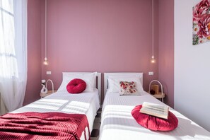 A bedroom with two beds, each with a white headboard, a red pillow, and a book on the right bed. There are bedside tables with lamps, a window with sheer curtains, and a wall with a floral painting.