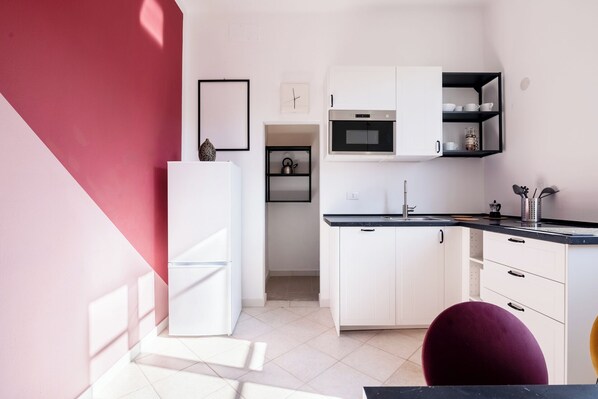 A compact kitchen with white cabinets, a microwave, and a sink. There is a small shelf with kitchenware and a decorative vase on top of the refrigerator.
