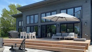 A modern two-story house with a large deck, outdoor seating, and a grill.