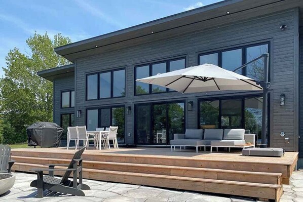 A modern two-story house with a large deck, outdoor seating, and a grill.