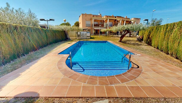 Ein rechteckiger Swimmingpool mit Fliesenränder, umgeben von einem niedrigen Hecken und einem Gebäude mit mehreren Balkonen im Hintergrund.