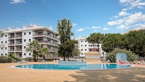 Un área de piscina con sillones de descanso, una fuente y un edificio de varios pisos con balcones al fondo.