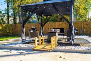 Ein überdachter Sitzbereich im Freien mit Tisch und Stühlen, einer Holzterrasse und einem eingezäunten Garten.