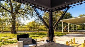 An outdoor patio with a table and chairs, a covered seating area, and a view of a fenced park with trees.
