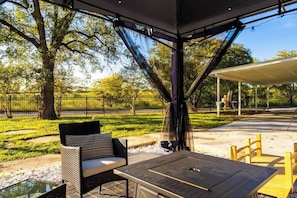 Eine Terrassensitzgelegenheit mit Tisch und Stühlen, ein überdachter Bereich zum Verweilen und Blick auf einen Park mit Bäumen, der durch eine Ziegelmauer begrenzt ist.