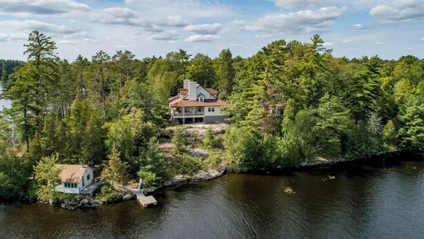 Una propiedad frente al lago con una casa principal y una cabaña más pequeña, rodeada de un bosque denso y un lago tranquilo.