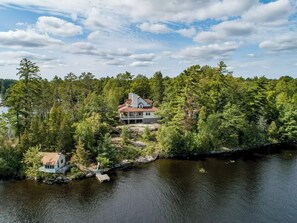 Una propiedad frente al lago con una casa principal y una cabaña más pequeña, rodeada de un bosque denso y un lago tranquilo.