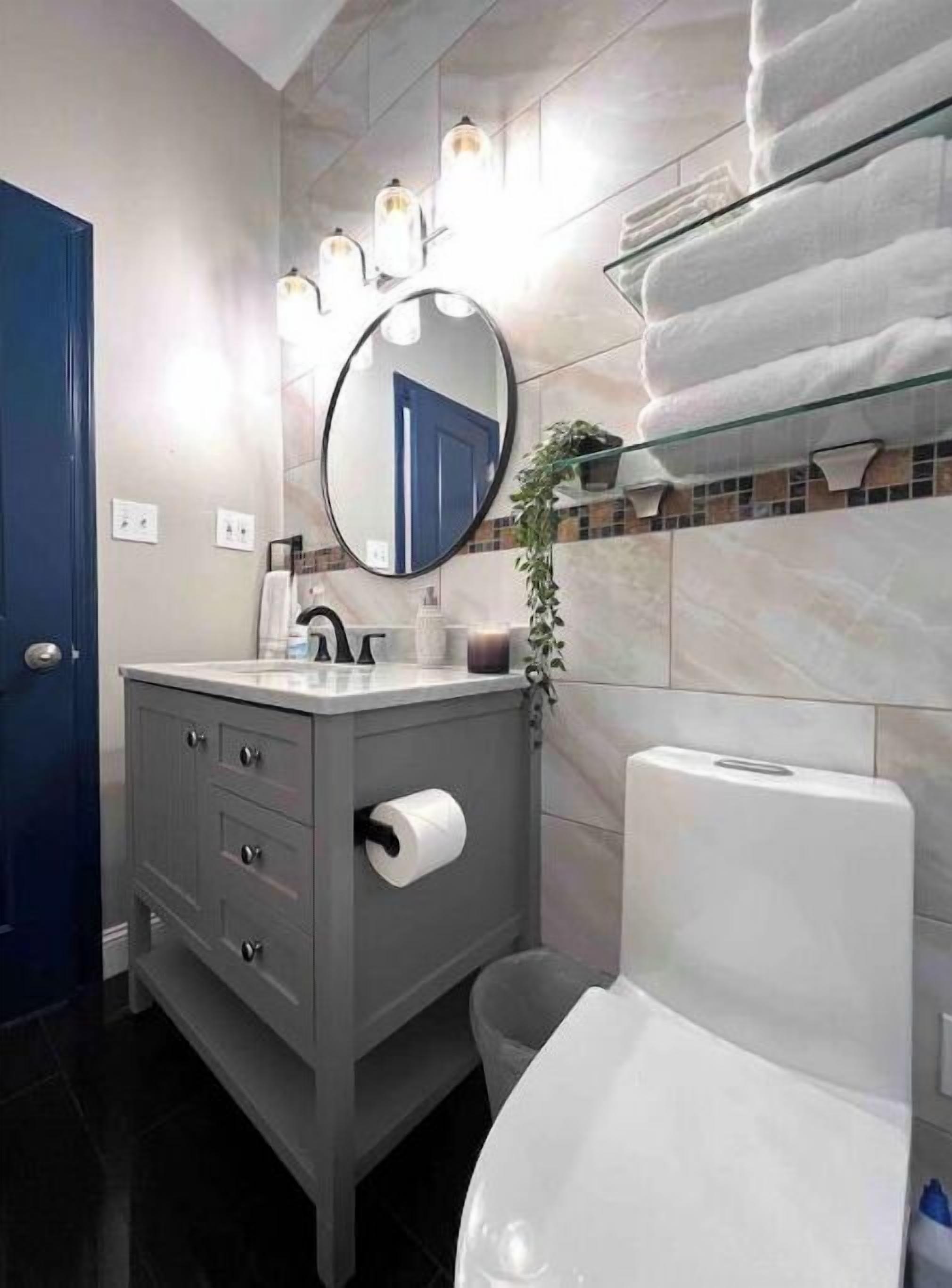 Une salle de bain avec un meuble-lavabo gris, un miroir rond, des toilettes et un porte-serviettes avec des serviettes pliées.