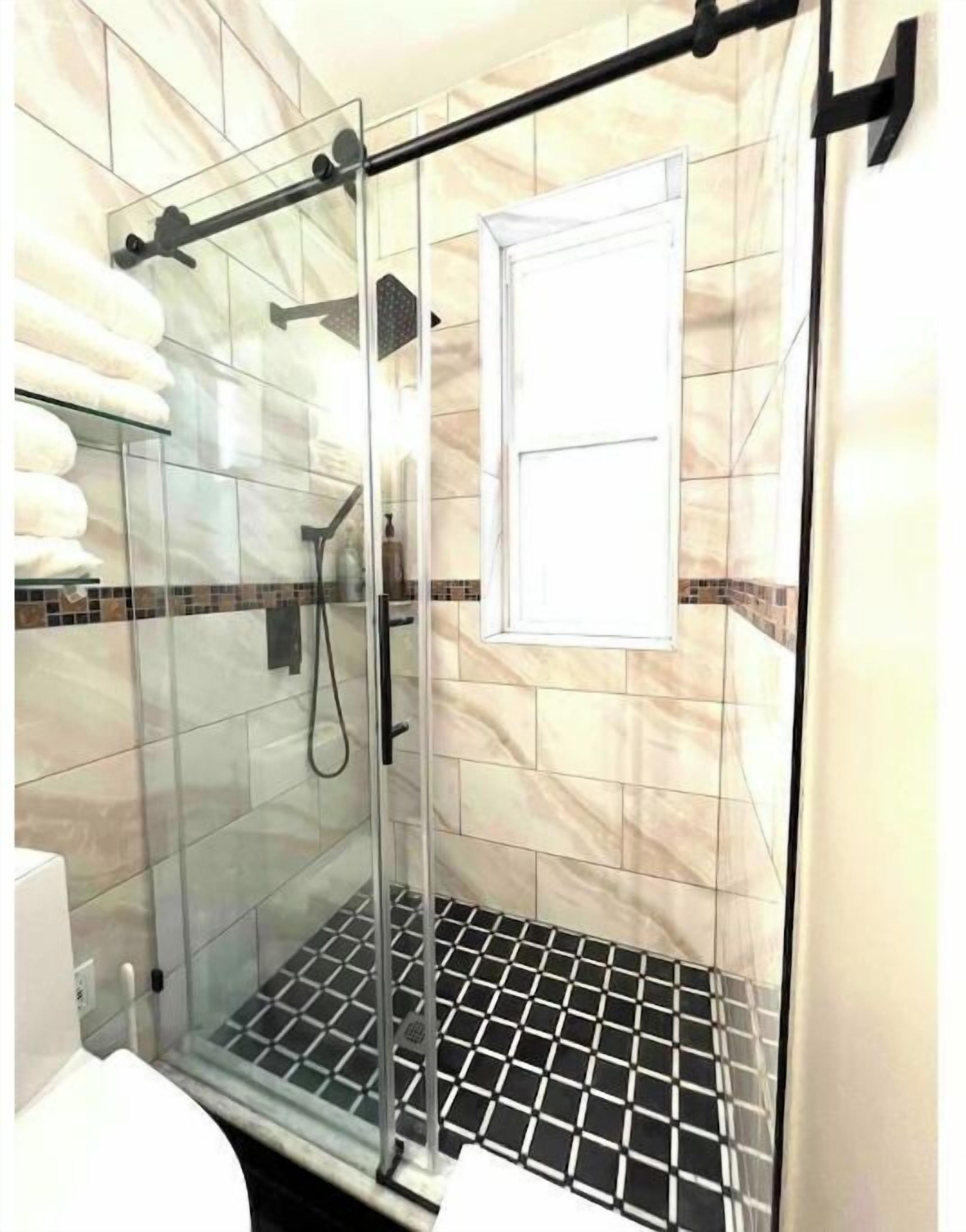 Une douche fermée par des parois de verre, avec un sol carrelé en noir et blanc, un porte-serviettes et une fenêtre à cadre blanc.