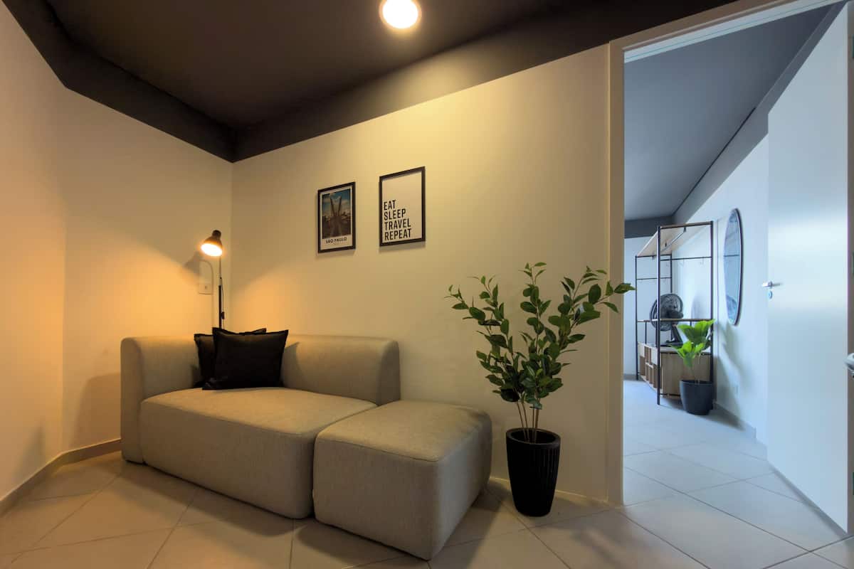 Ein modernes Wohnzimmer mit einem beigen Sofa, einer Stehlampe und einem Blumentopf. An der Wand hängen zwei gerahmte Bilder, und eine Treppe führt in einen anderen Raum.
