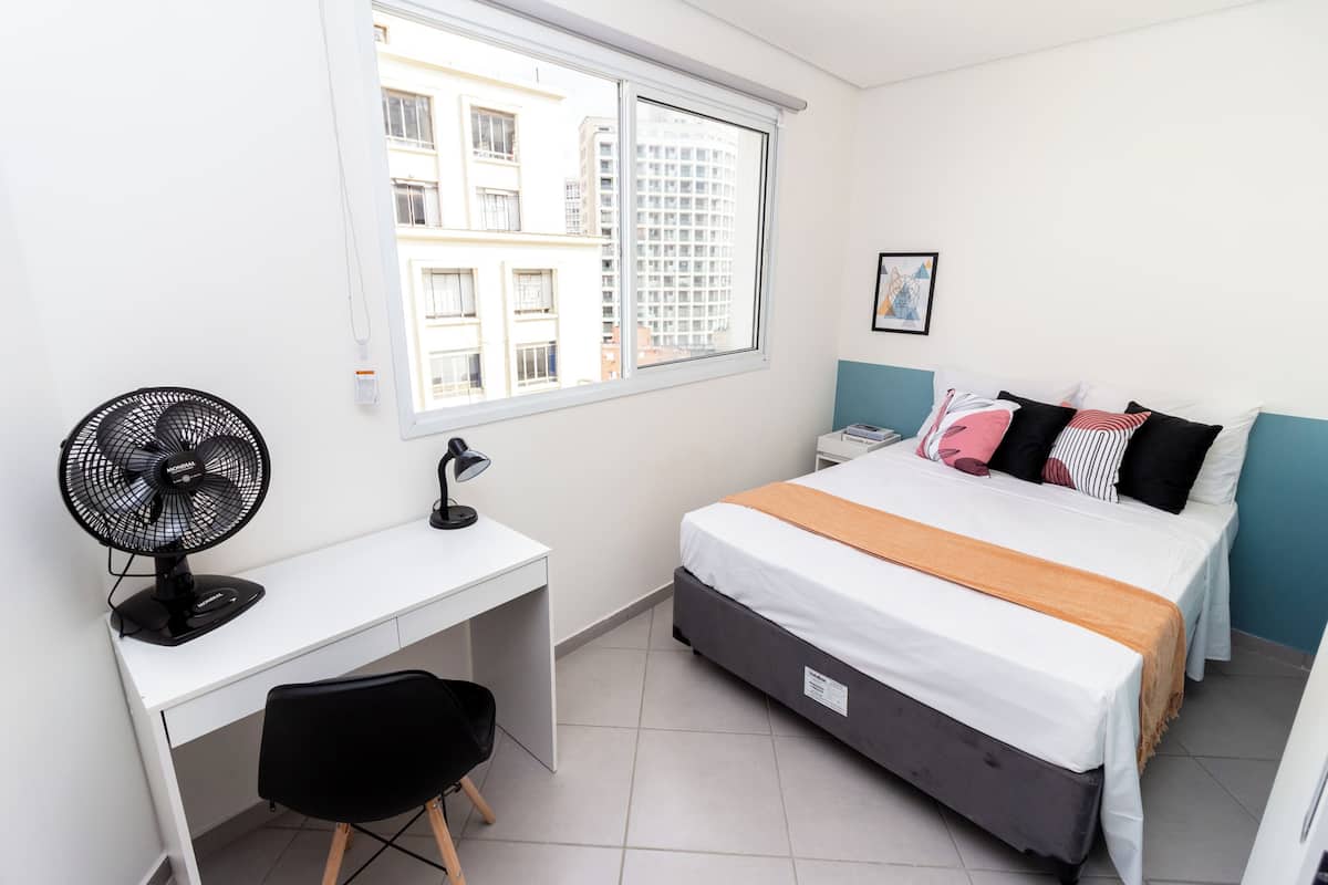Ein Schlafzimmer mit Bett, Schreibtisch, Stuhl, Standventilator und einem Fenster mit Blick auf die Stadt.