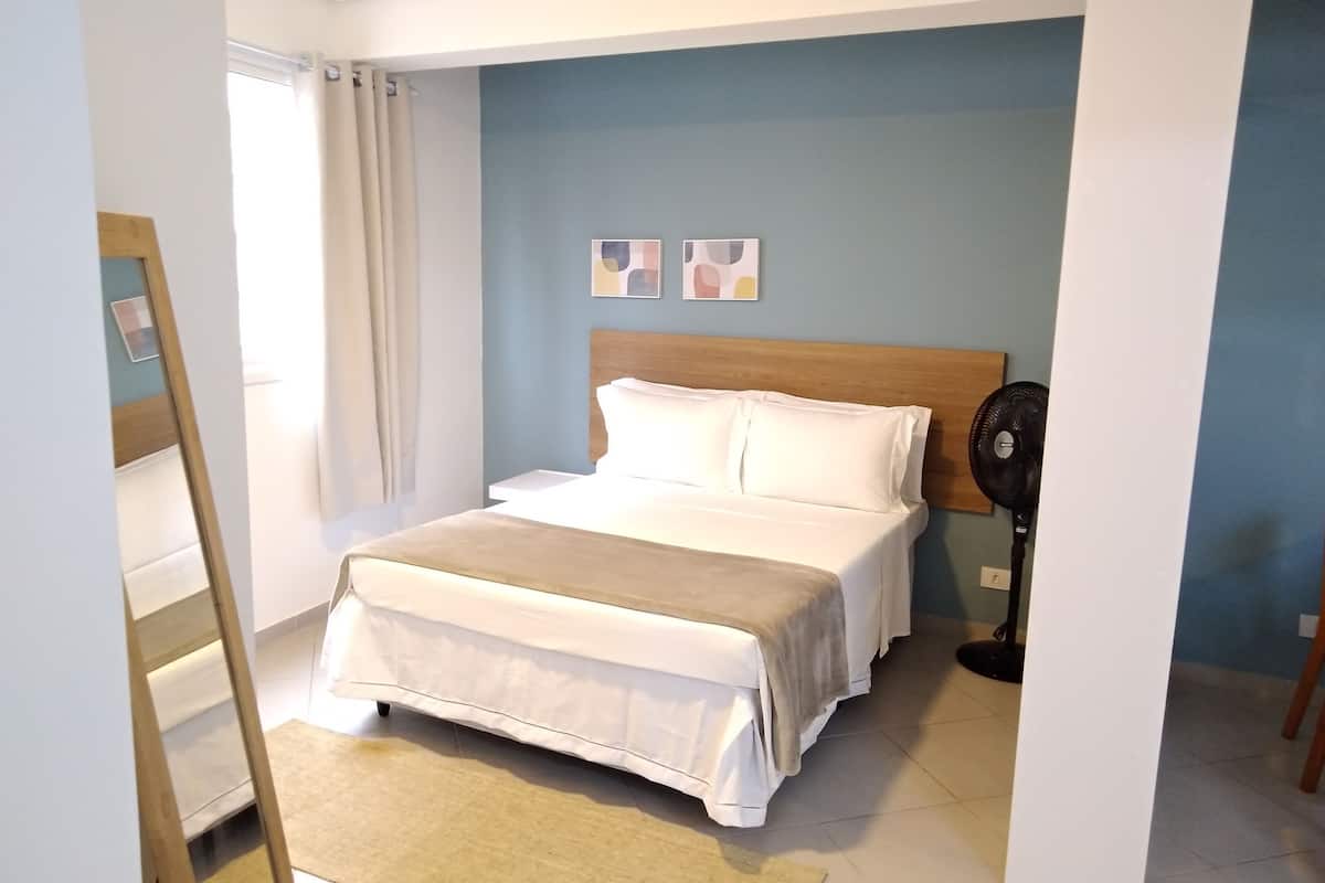 Ein ordentlich bezogenes Bett in einem Zimmer mit blauer Wand, einem Holzkopfbrett und einem Standventilator.