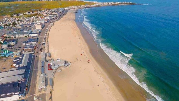 Vue aérienne d’une ville côtière avec une longue plage de sable, une mer bleue et limpide, et un mélange de bâtiments résidentiels et commerciaux le long de la côte.