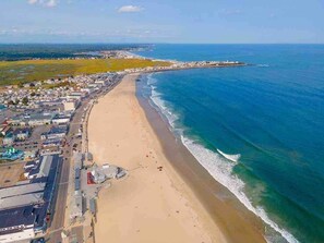 An aerial view of a coastal town with a long sandy beach, a clear blue sea, and a mix of residential and commercial buildings along the shore. - Beachside Getaway - Steps to the Beach (Hampton)
