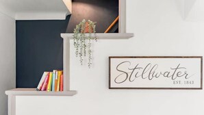 Uma estante com livros coloridos, uma placa emoldurada com a inscrição "Stillwater EST. 1843" e uma planta pendente.